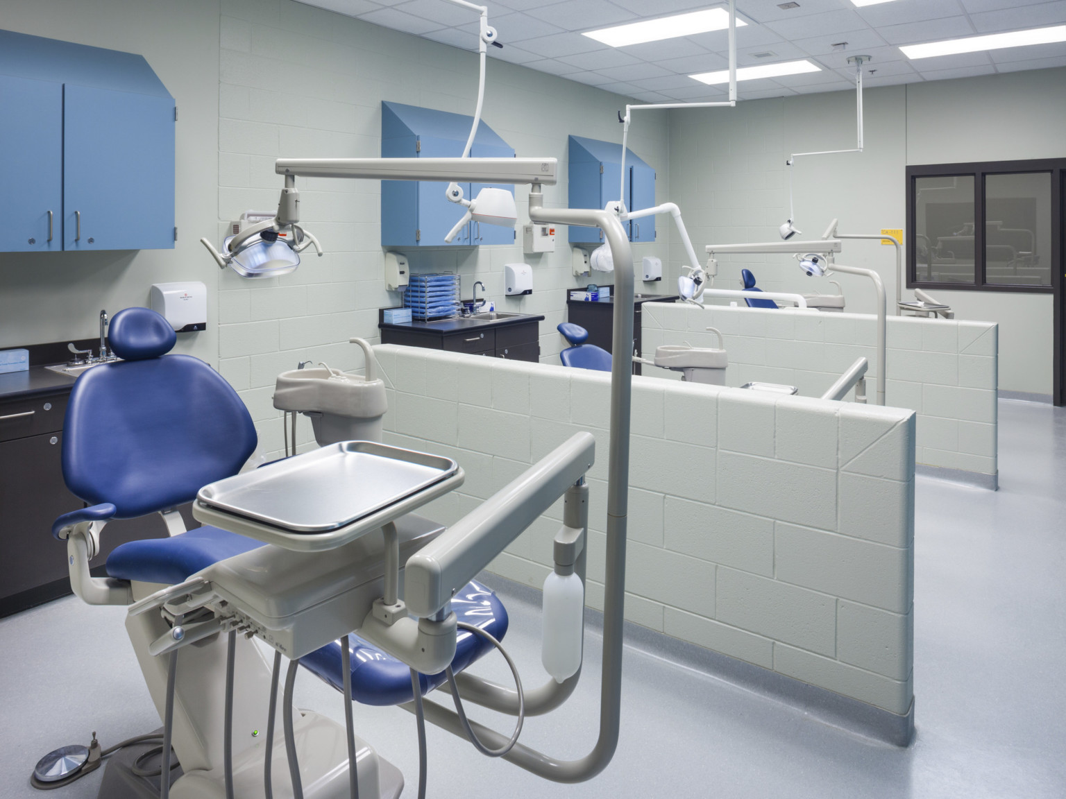 3 dental exam chairs separated by concrete block half walls. Back wall has blue cabinets behind each chair