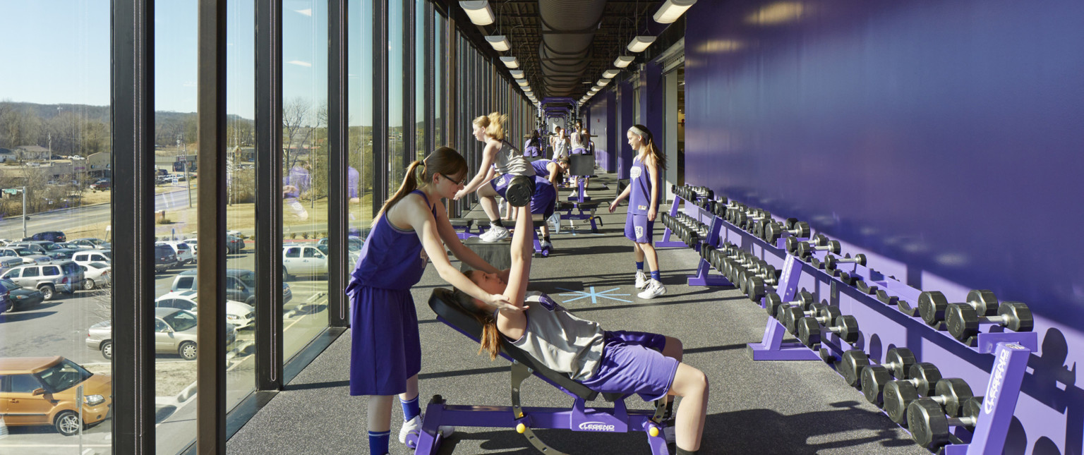 Hallway with purple wall right and wall of windows to the left. Girls use weights and training equipment below black ceiling