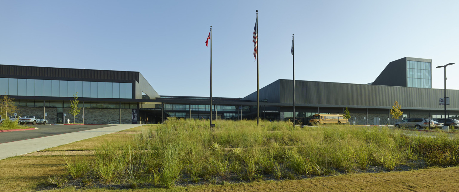 View from across lawn with 3 flags. Stone lower level with glass entry below black 2nd floor with large seamless windows