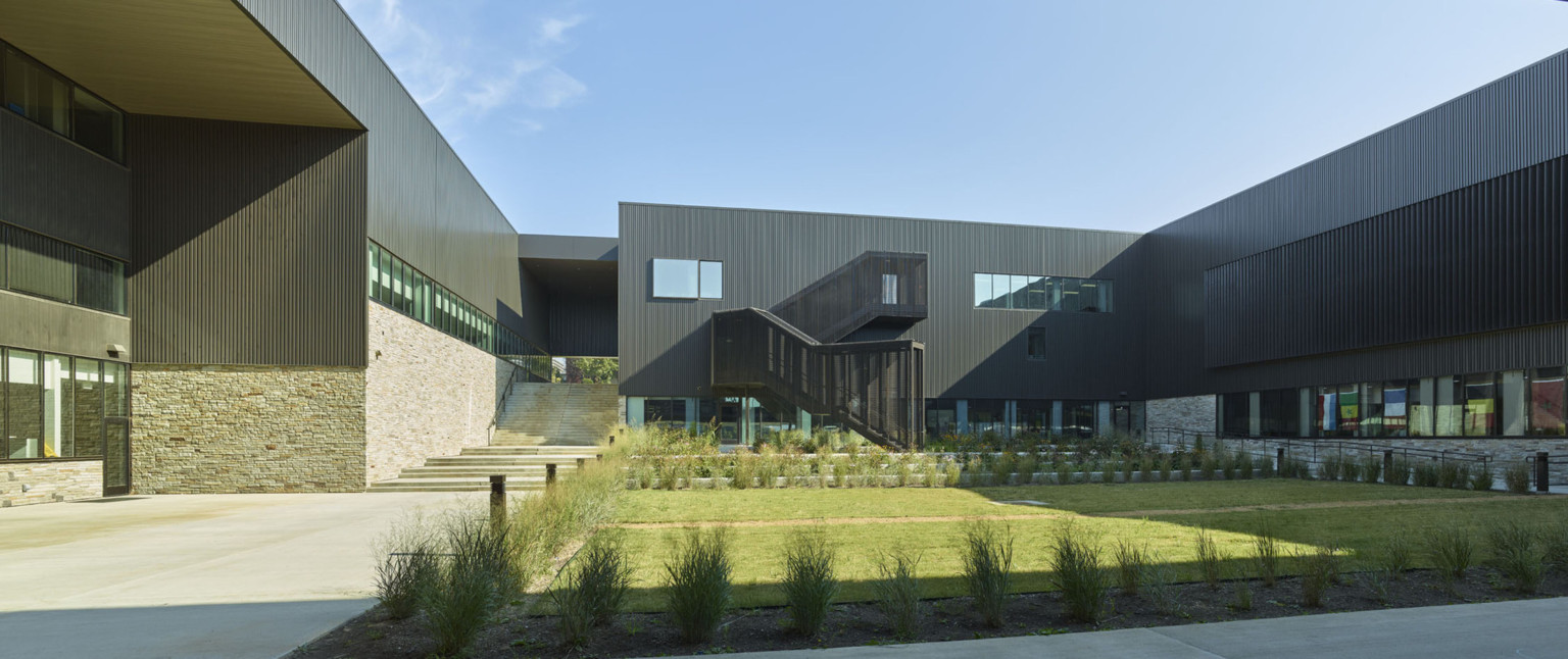 Courtyard surrounded by building with stone lower level and steps to left. Black paneled upper walls with sheltered stairwell