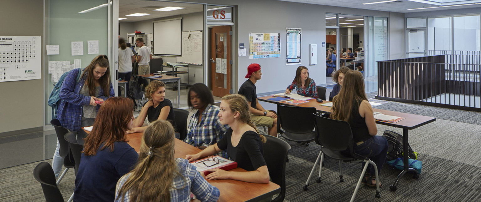 Students sit at long tables in grey corridor. Classrooms are left, with transparent wall looking in. Right, a black bannister