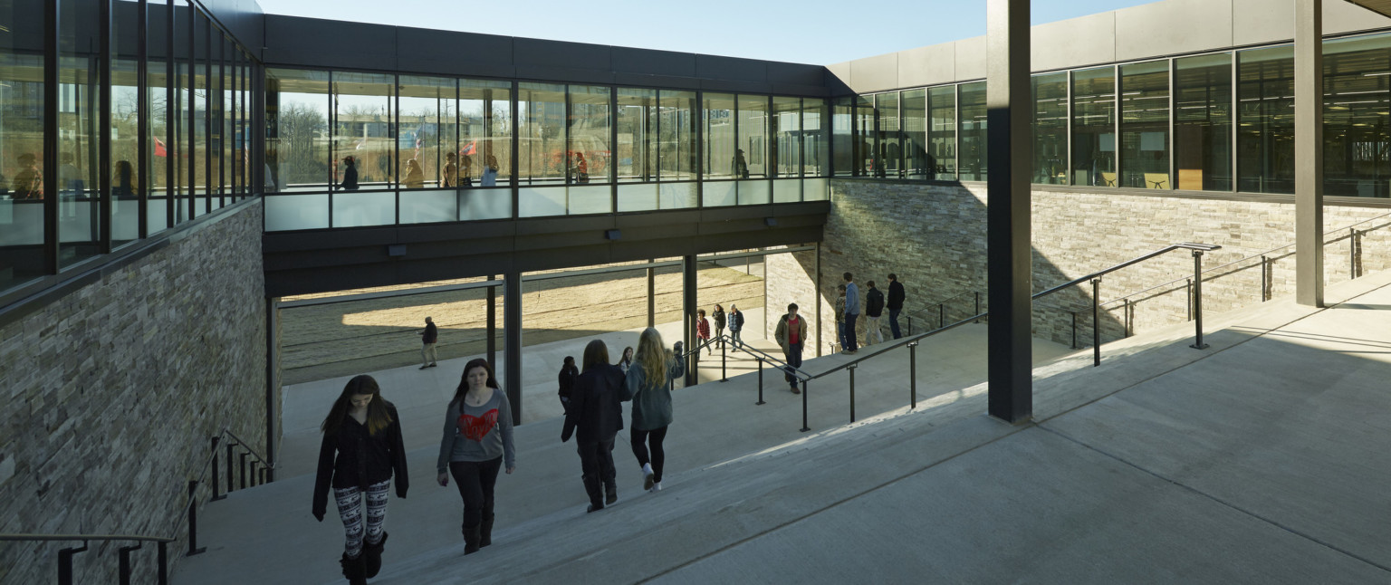 Outdoor stairwell viewed from covered path above. Walls on either side with stone base below glass lined hallway on 3 sides