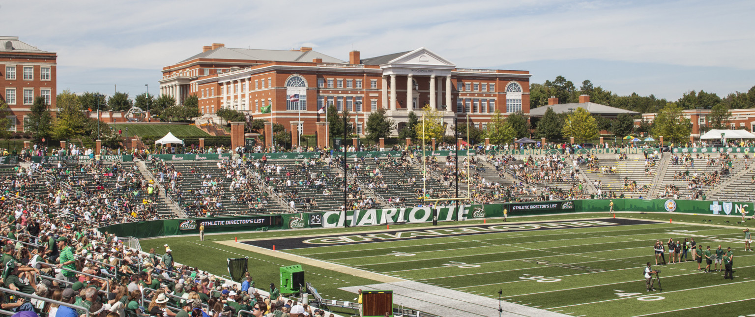 View from stands of end zone with Charlotte written across. Bleacher seats behind green wall, brick and metal fence at back