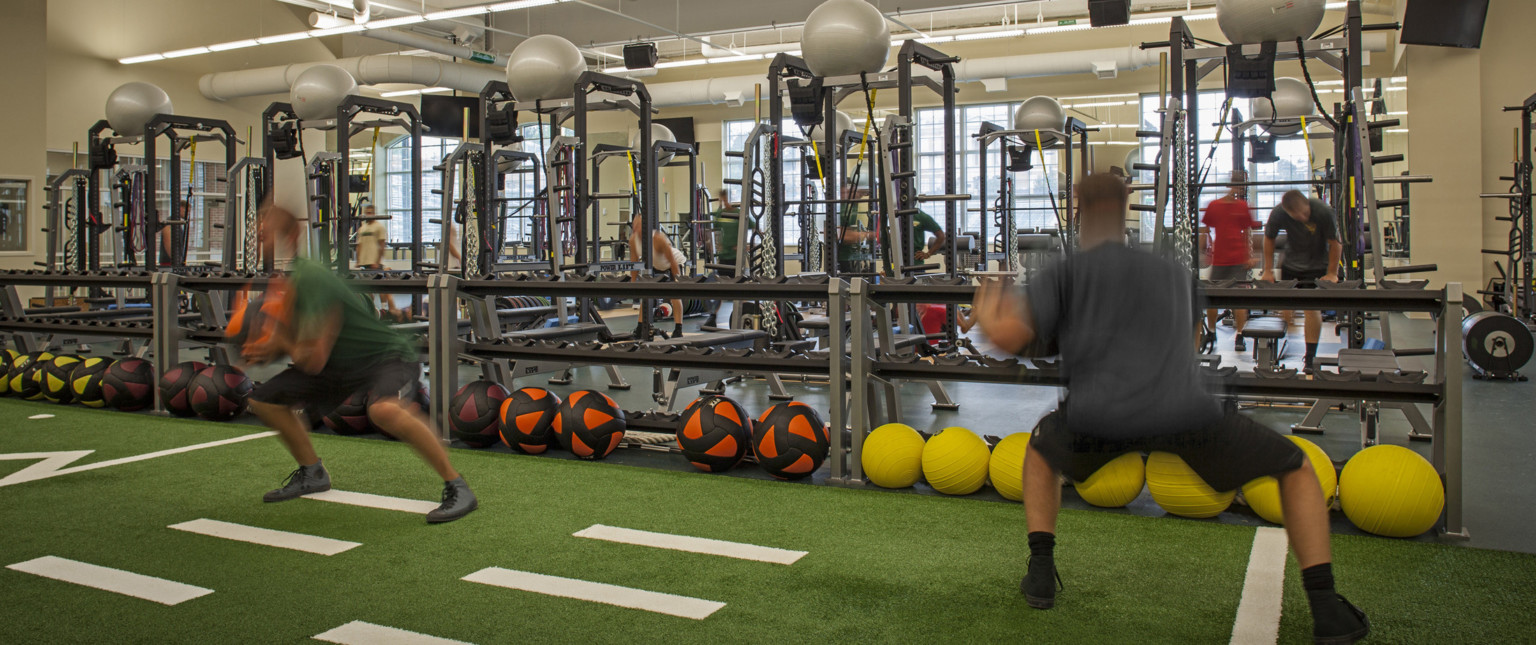 Turf floor space with yardage marked lined with half wall weight shelves in front of training equipment in cream color room