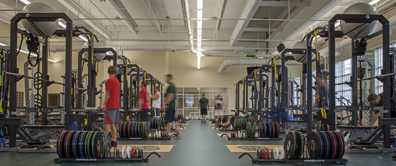 Weight room in training facility with equipment. Window lined cream colored walls under white ceiling with exposed ductwork