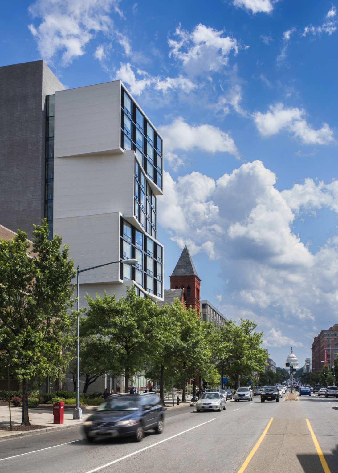 modern white building with glass front facade over a busy street with brick buildings and the capital dome in the background