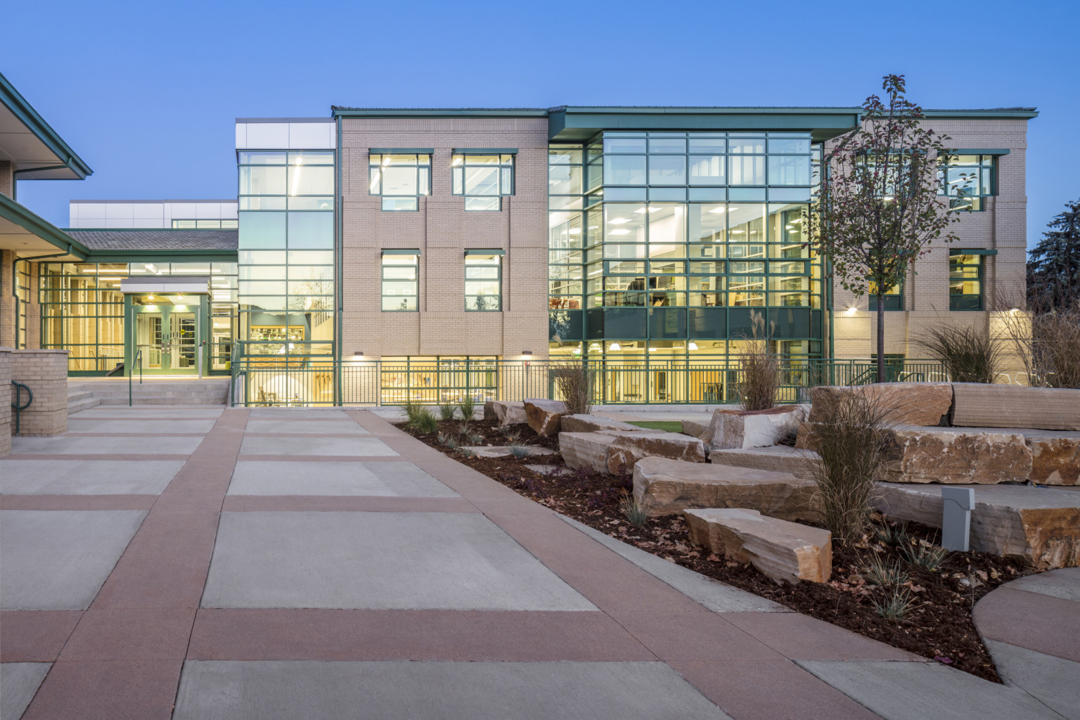 orthogonal brick and concrete walkway next to rocky landscaping in front of brick and three story illuminated glass facade