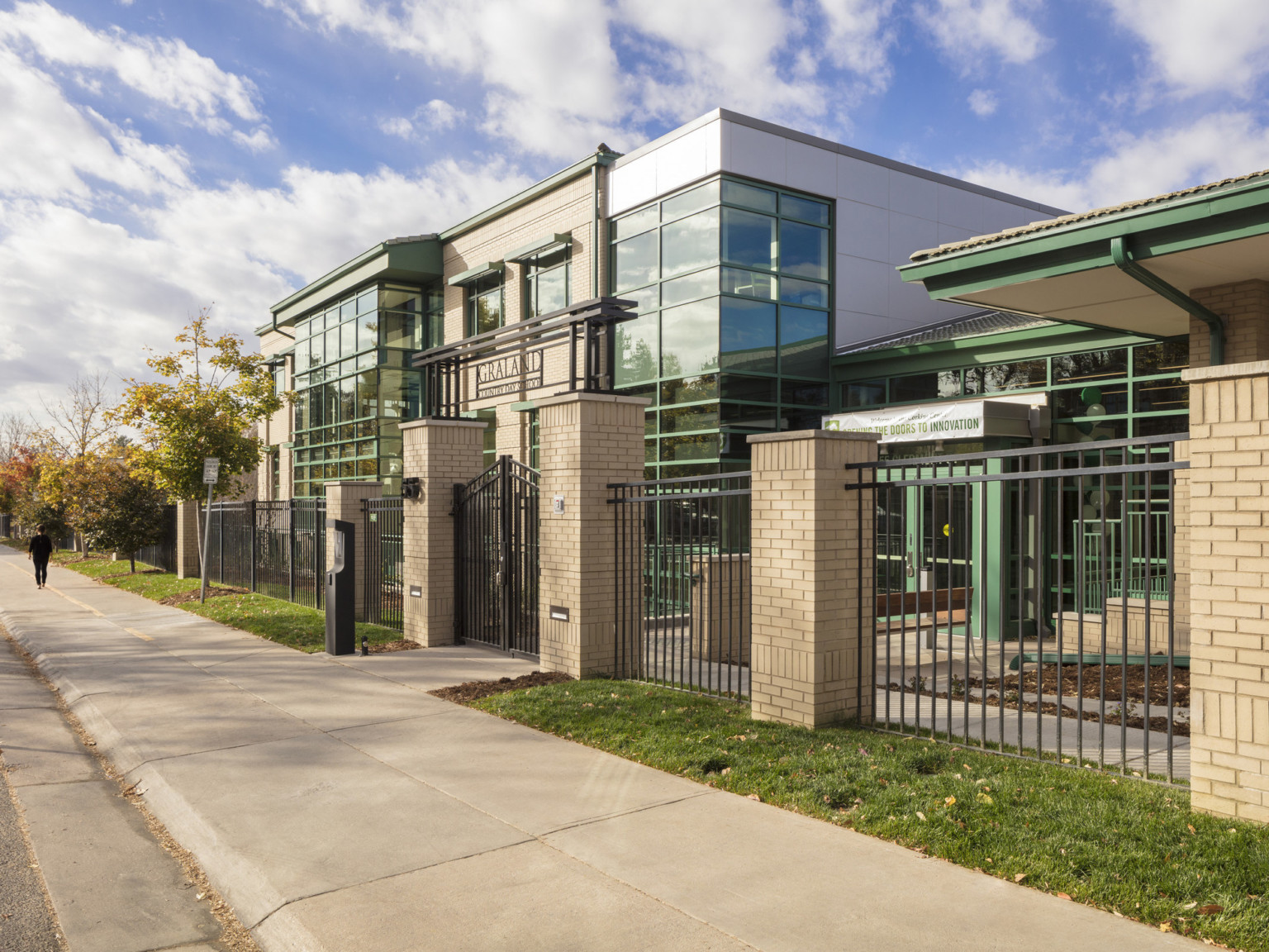 concrete sidewalk in front of brick and wrought iron fencing in front of a building with alternating glass and steel facade