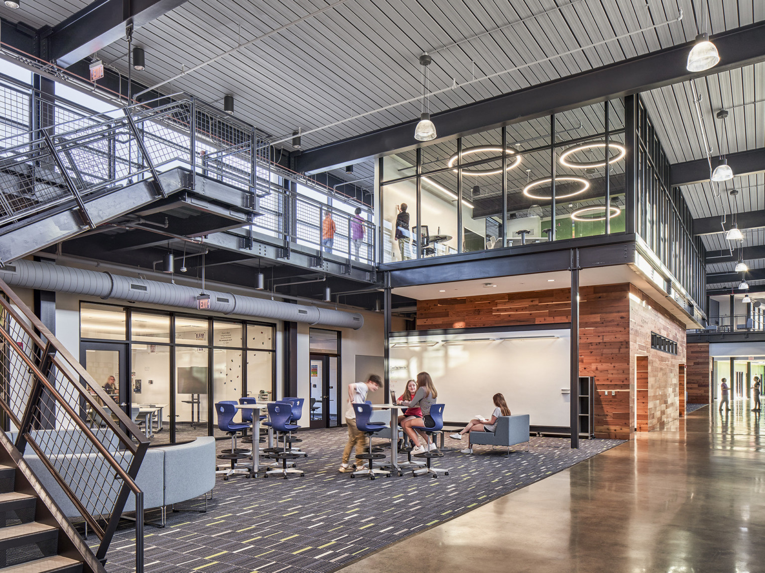 wide view of middle school collaboration area standing height tables, views to adjacent classrooms through glass doors, and a second-floor meeting room visible