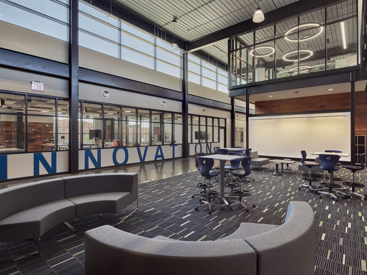 Innovation written on the far wall of a collaboration area at a middle school with curved couches, standing tables, and a large whiteboard beneath a visible second-floor lab