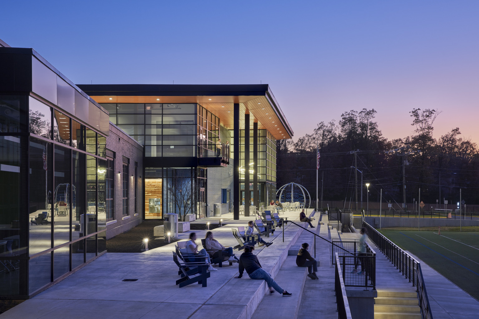 students relaxing on cement patio outside a modern school with glass walls, wood overhang held up with metal columns