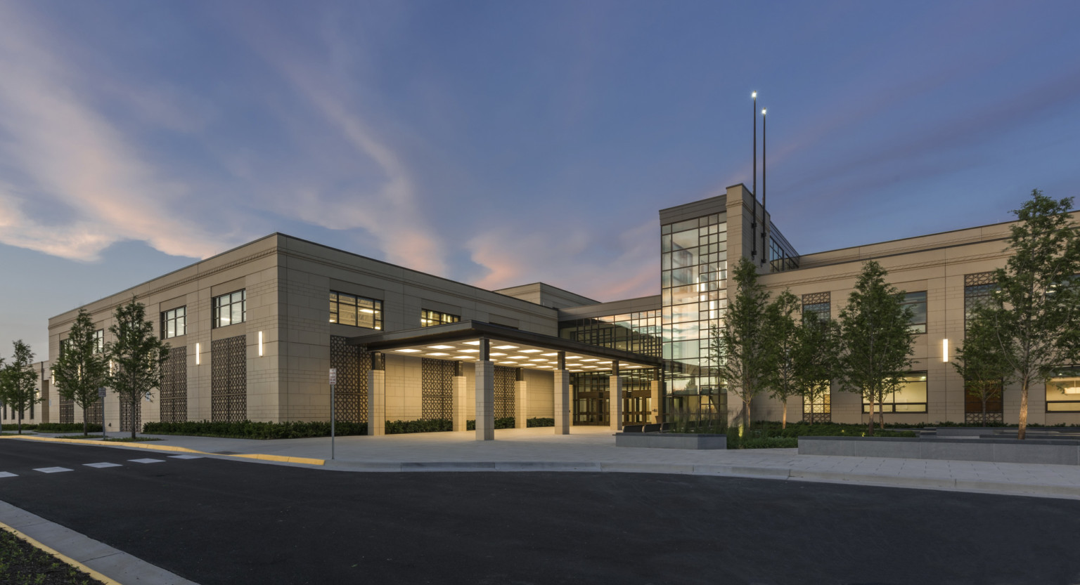 evening view of a precast concrete façade with an illuminated canopy entrance, a double-height glass atrium, and middle eastern inspired façade design