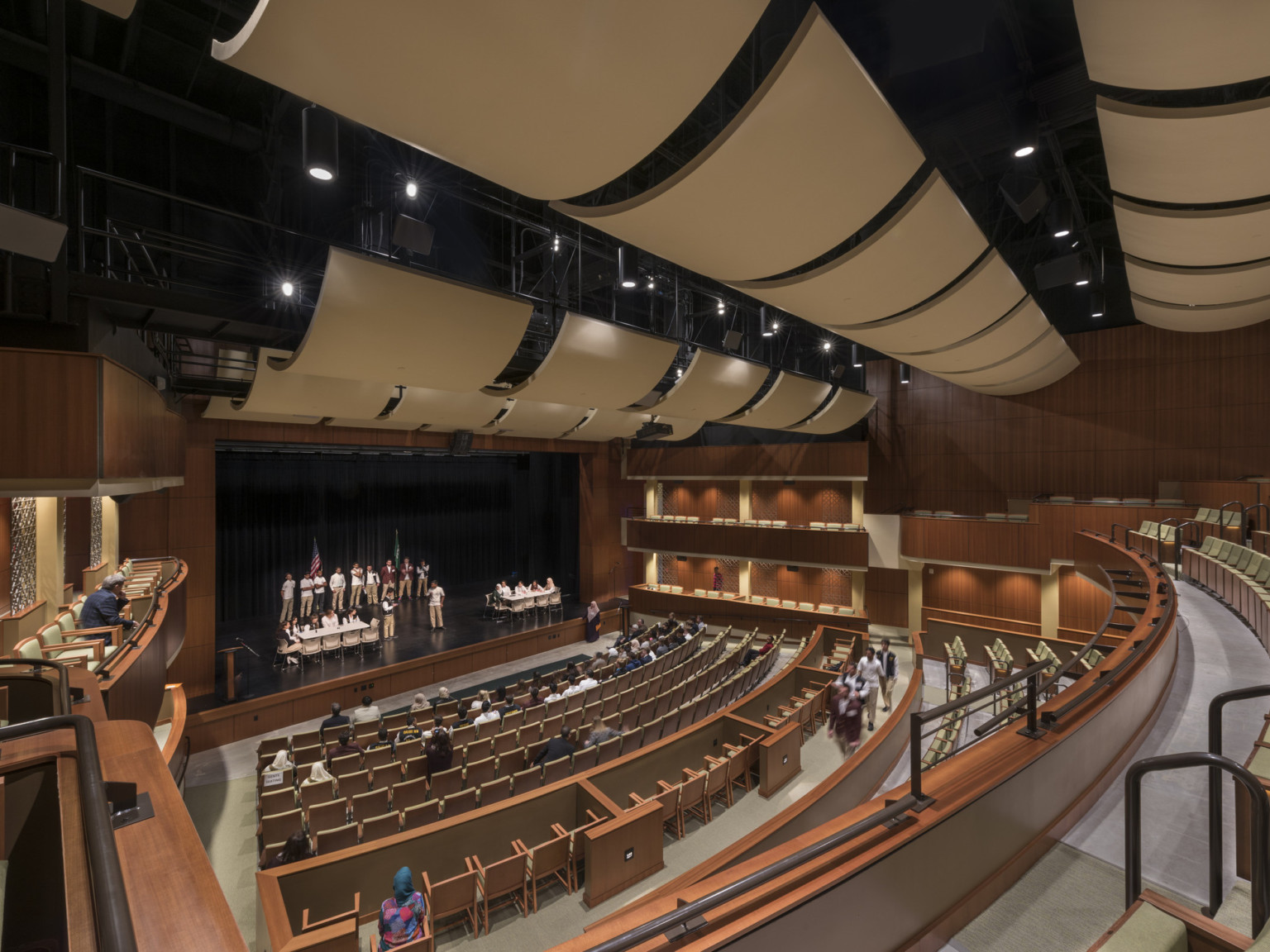 school theater with warm wood seating, box seating along the sides, beige acoustic ceiling panels, and black railings