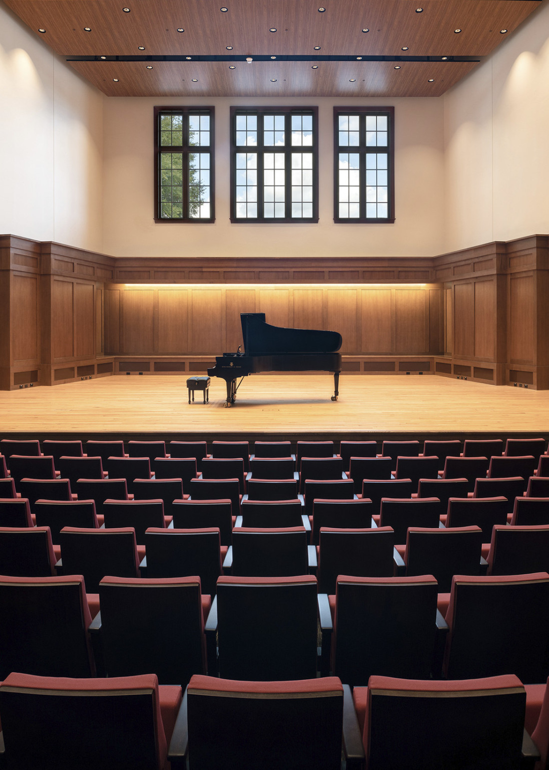 the backs of fixed theatrical seating facing a wooden recital stage with a black grand piano beneath three large windows