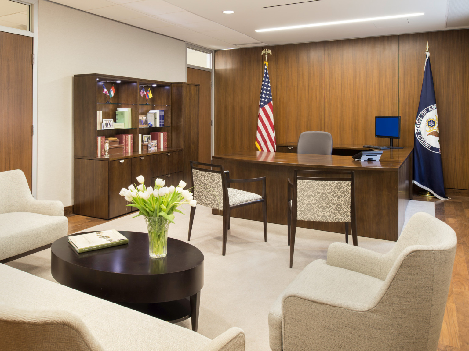 Wood desk, door, and rear accent wall. Left wall and carpet are white. White chairs with coffee table front, flags frame desk