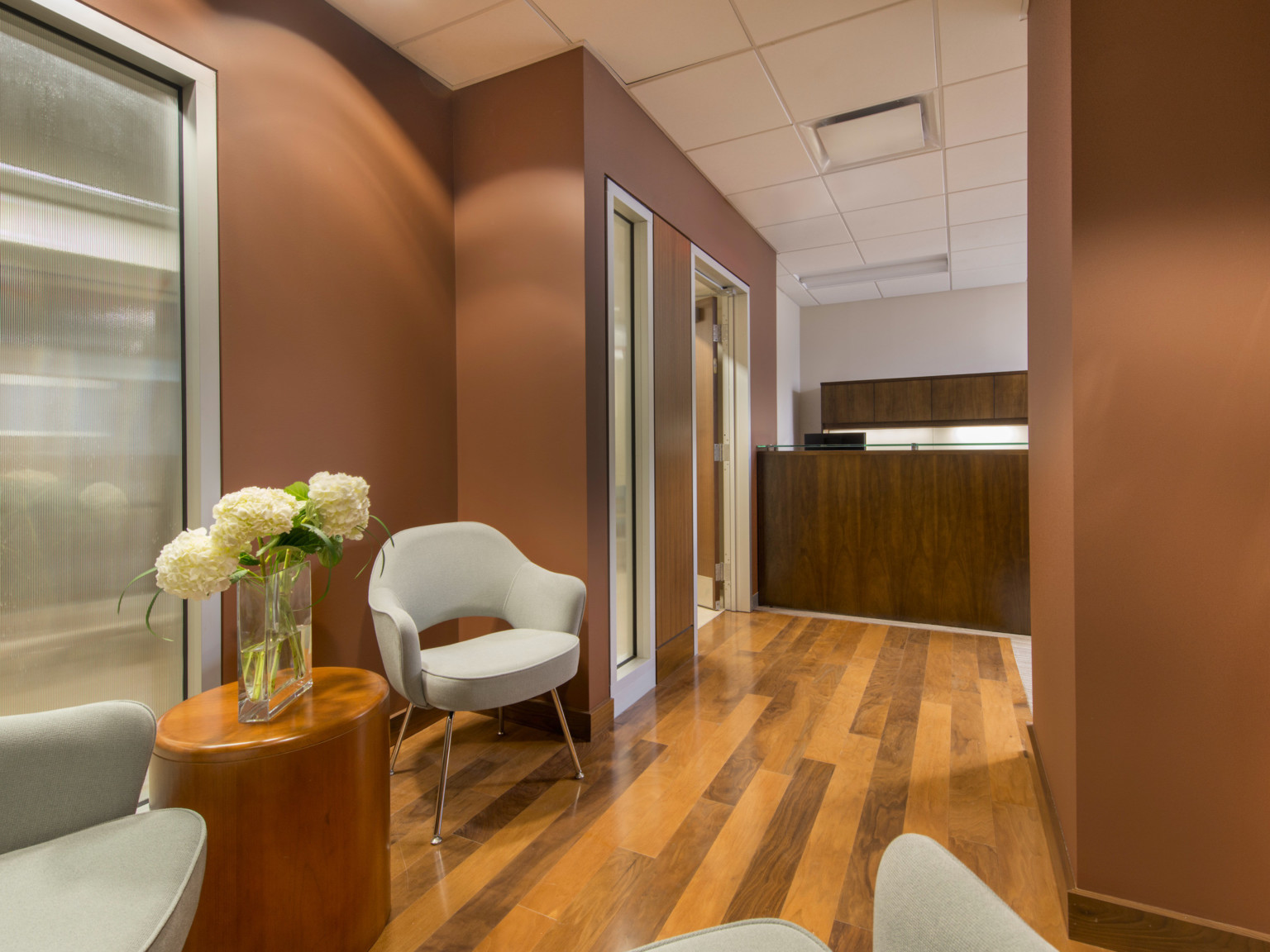 Hardwood floors below grey arm chairs with flowers on side table. Brown walls with translucent windows lead back to wood desk