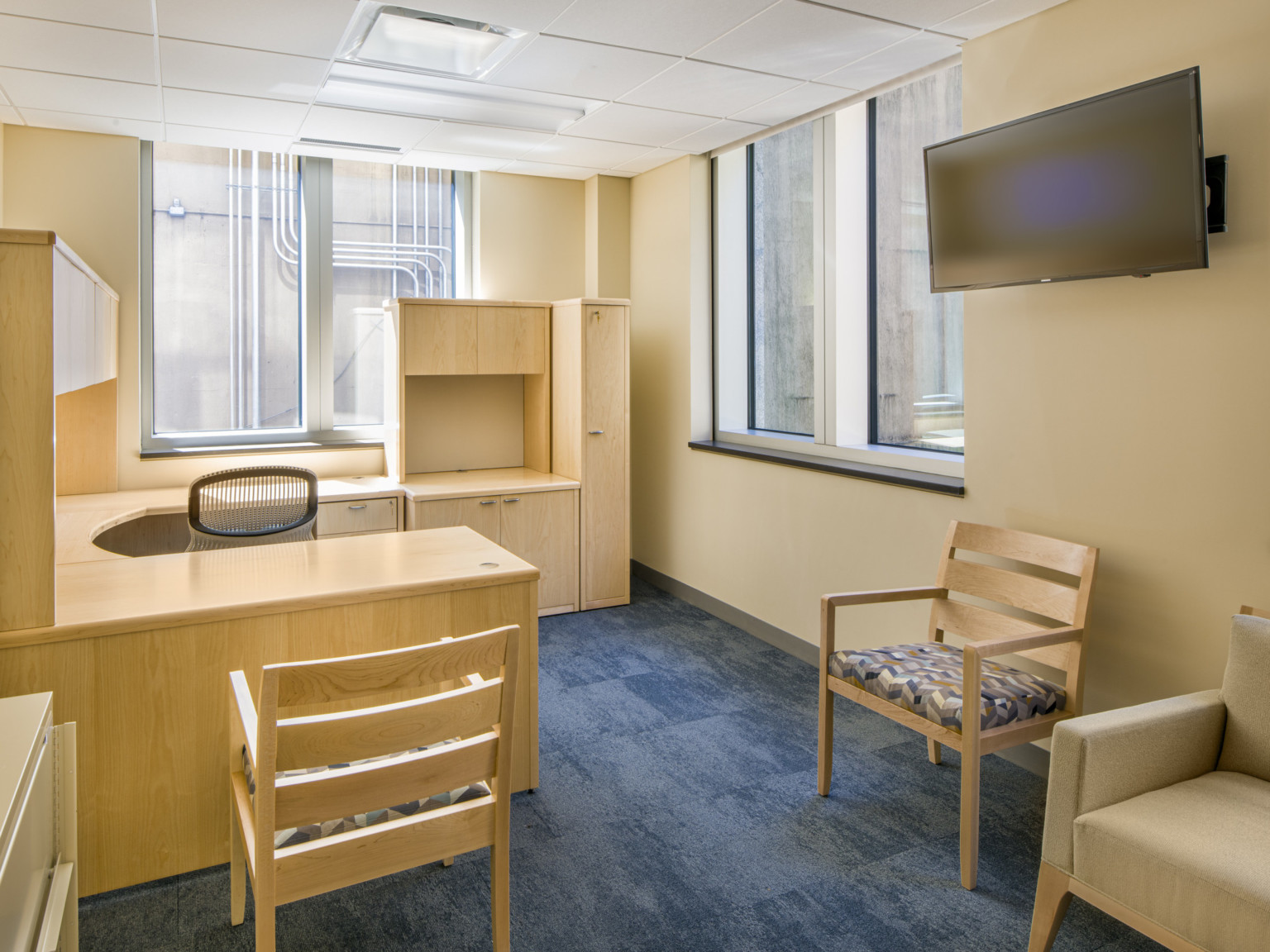 Office with cream walls and white ceiling, light wood wrap around desk and matching chairs. Windows on right and back wall