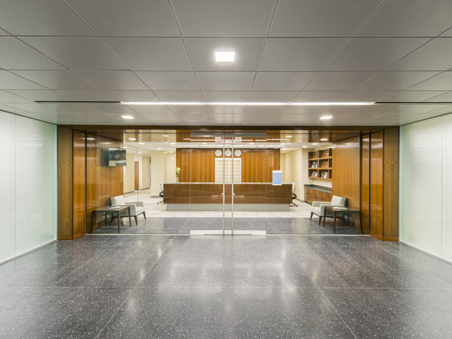 Hallway with reception area behind glass double doors. Hall is white with grey flooring, reception has wood walls and desk