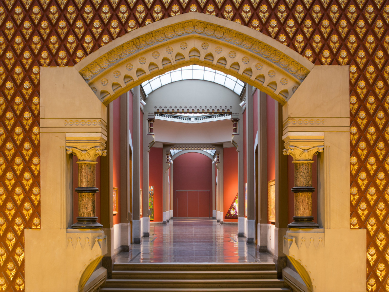 Corridor entry with sculpted stone archway with banded Corinthian columns. The hall is painted red with skylights above