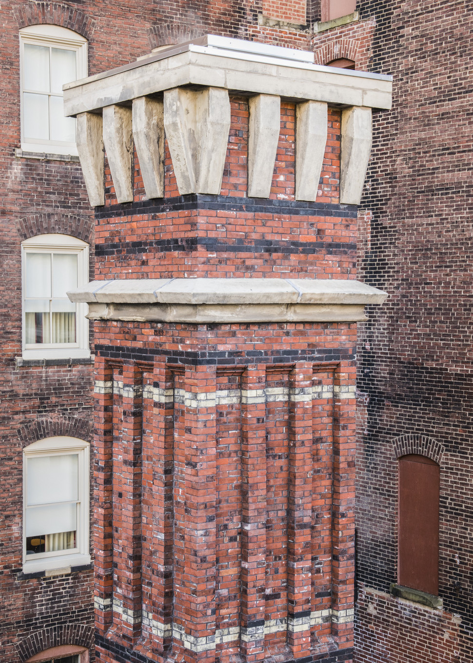 Red brick chimney with black and white horizontal stripes and light colored stone details at top in front of a brick wall