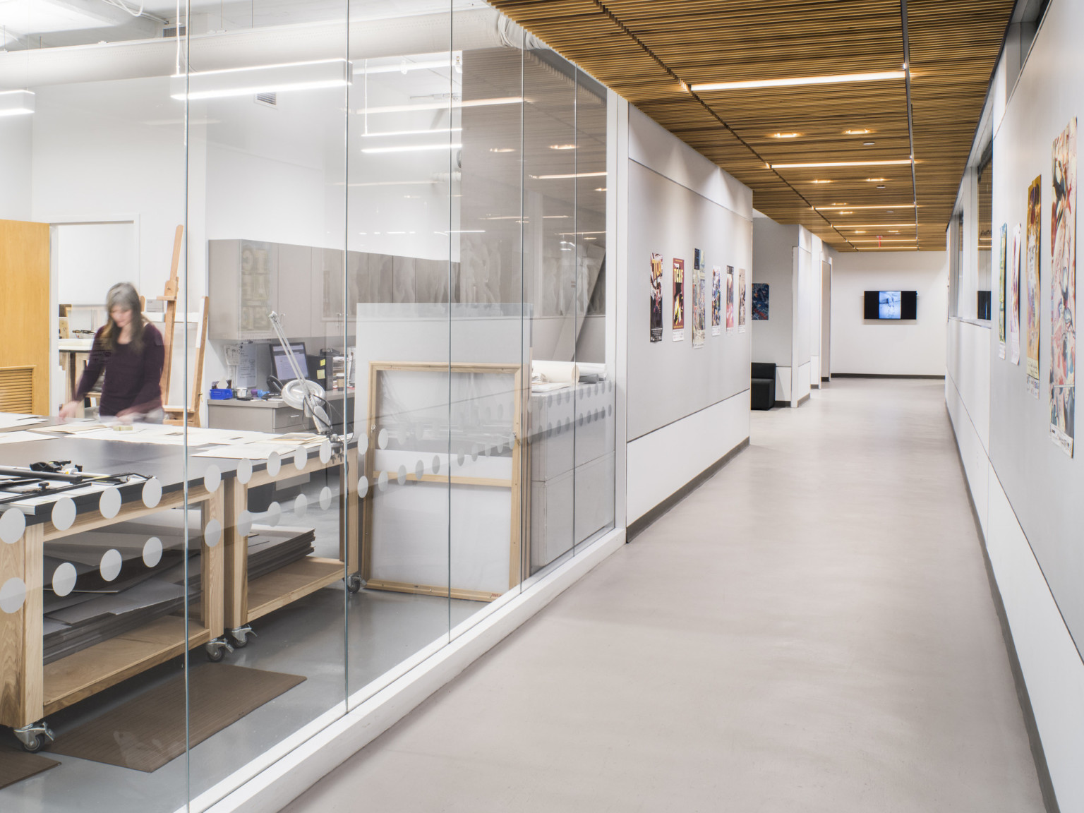 A hallway with a glass walled room, in which a woman works with papers in front of her at one of the tables