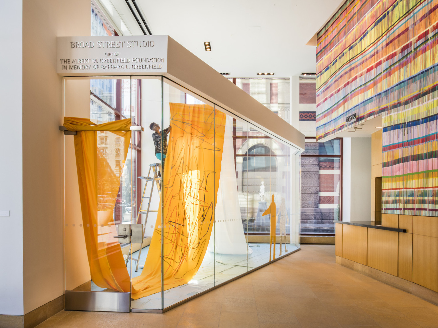 Glass exhibit booth in front of window to the left, right wall has textural multicolored striped wallpaper above wood base