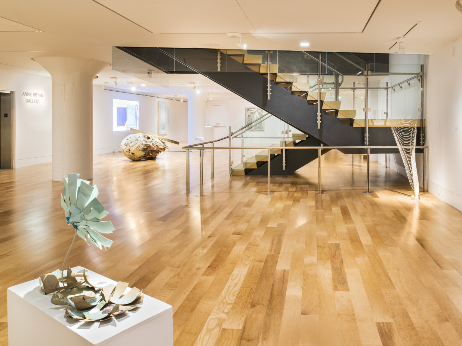 White art gallery with light color hardwood flooring and stairs with black base leading upstairs. Art is on white pedestal