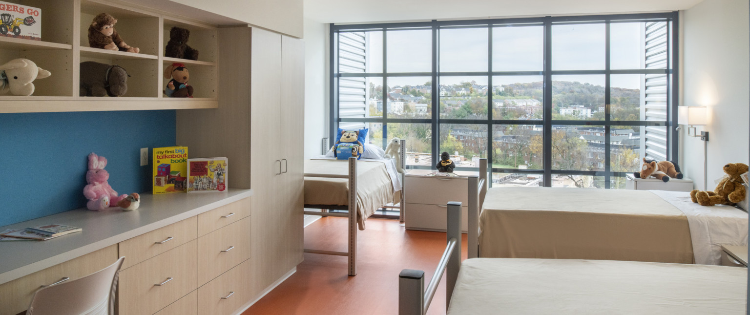 Interior of children's bedroom facing geometric window wall. 3 beds, closets and counter space left with books and toys