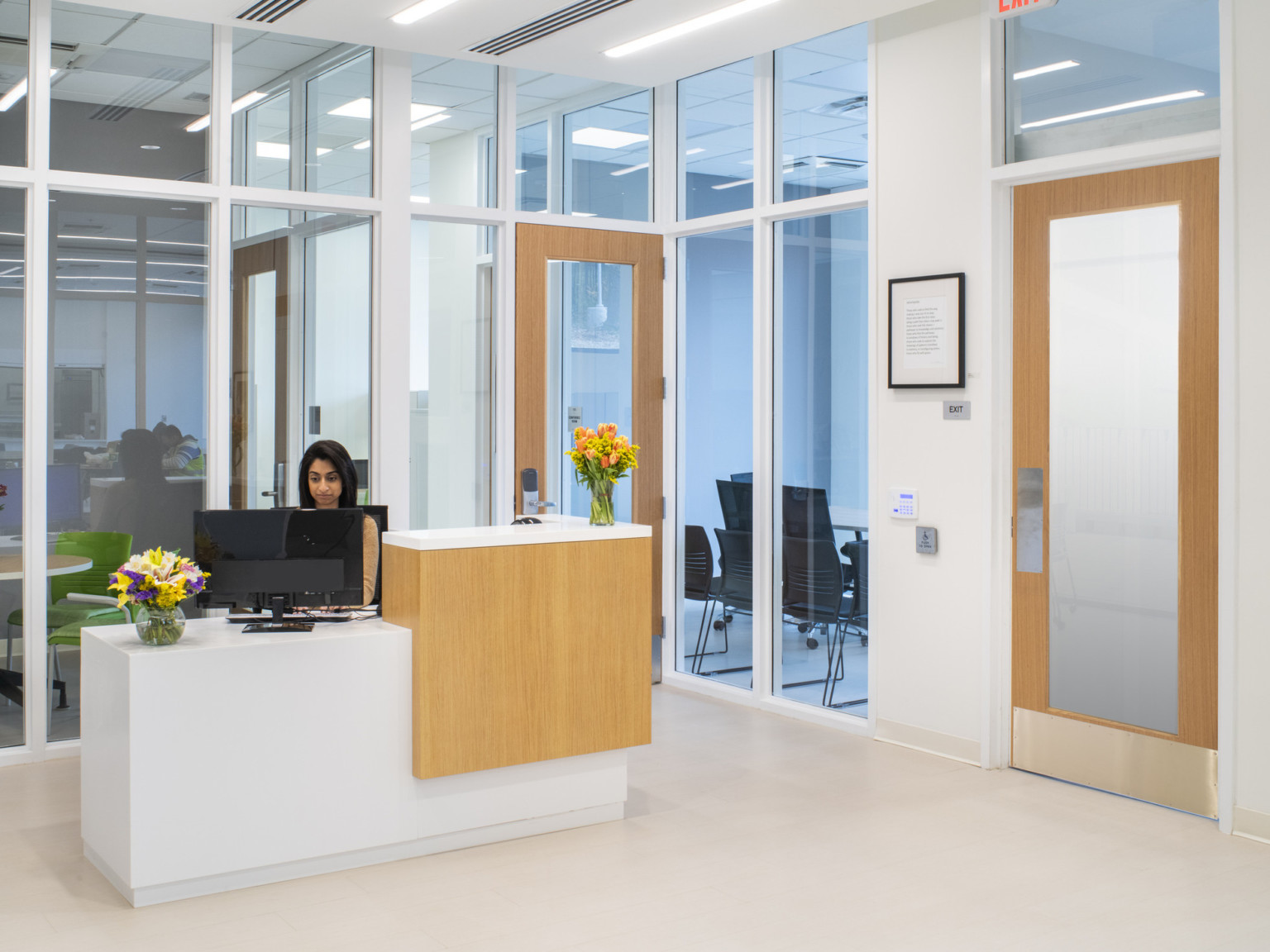 Woman sitting at white desk with wood accents which match office interior. Windows separate office and surrounding spaces