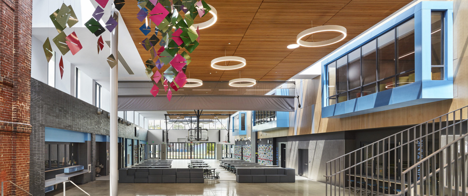 Double height atrium with exposed brick, left, and blue overhanging 2nd floor windows, right. Multicolor sculpture hangs left