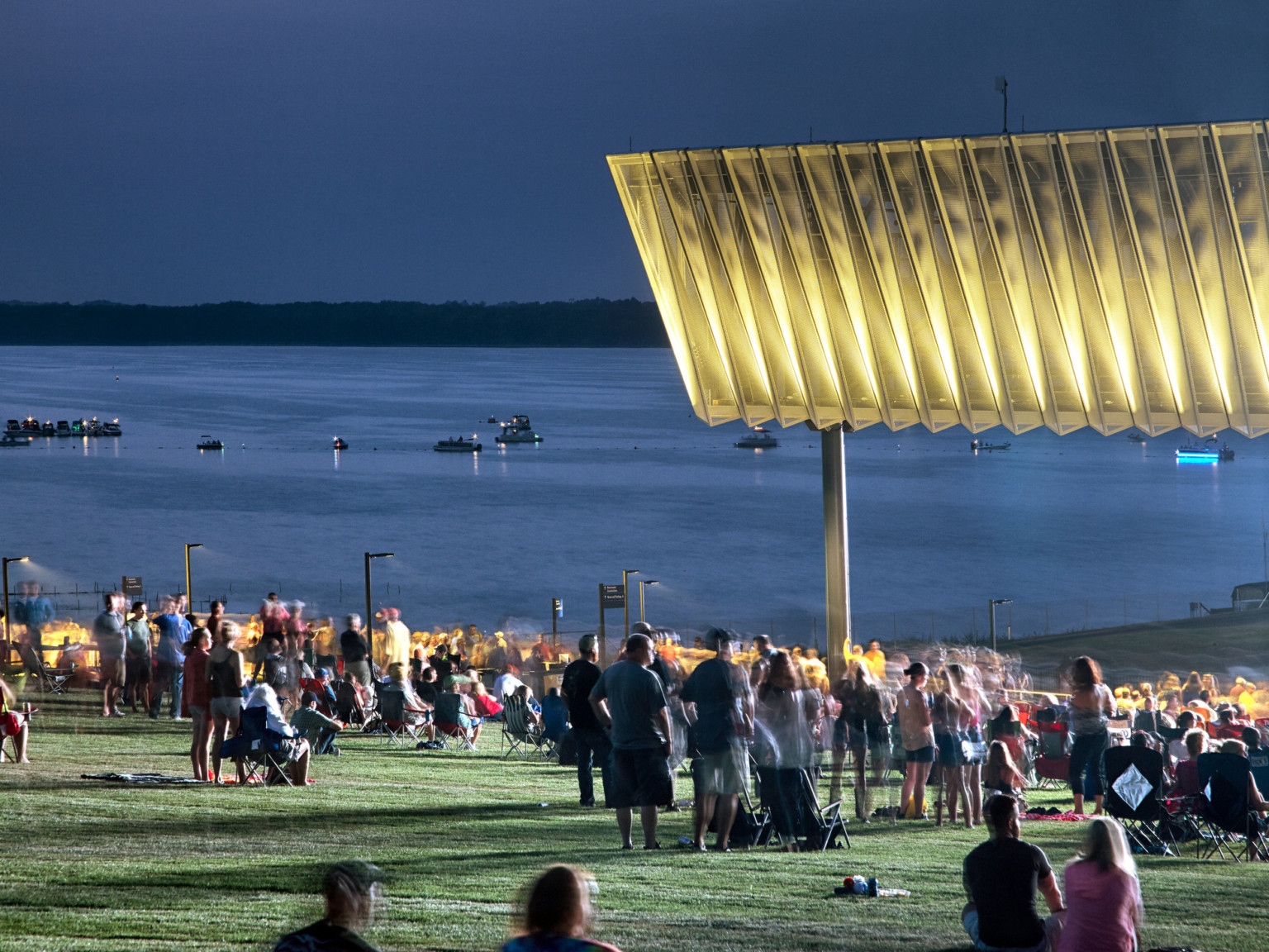 Crowd under illuminated fascia at night in front of lake with boats