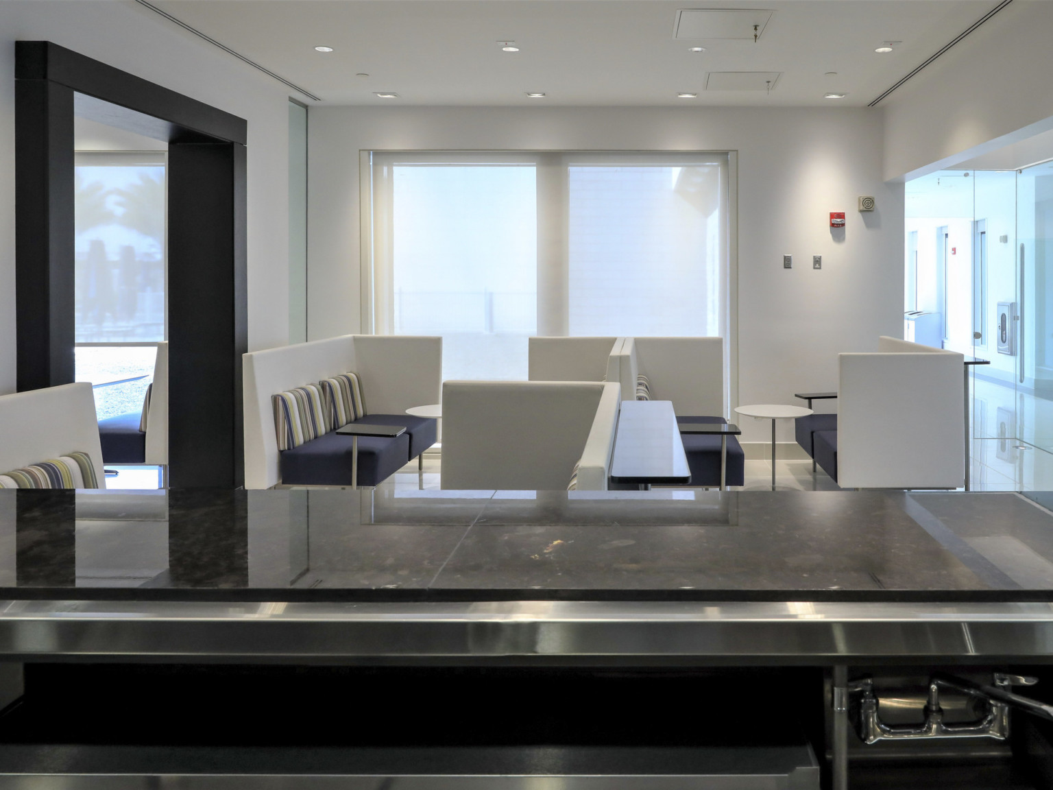 Couch seating area with tables in white room, seen from behind black counter top. Left is a black doorframe, right a hallway