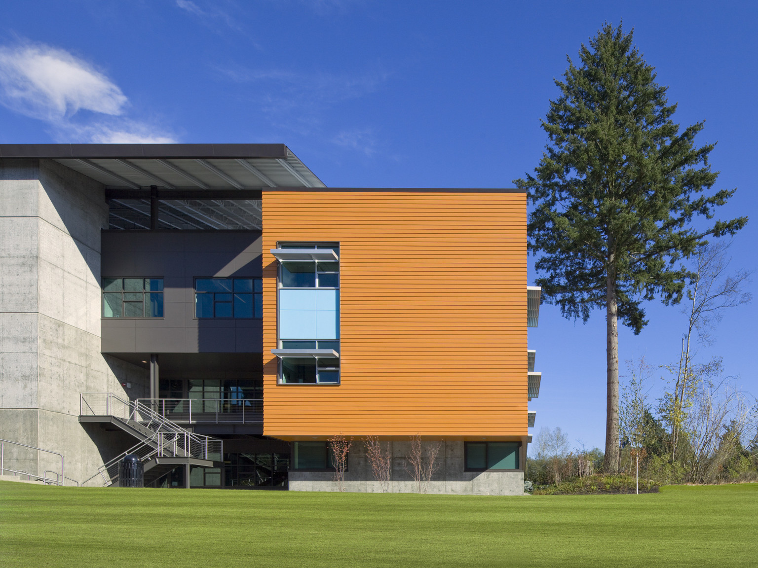 Concrete wall of building to left of recessed dark grey building section under large panel. Orange section connects at right