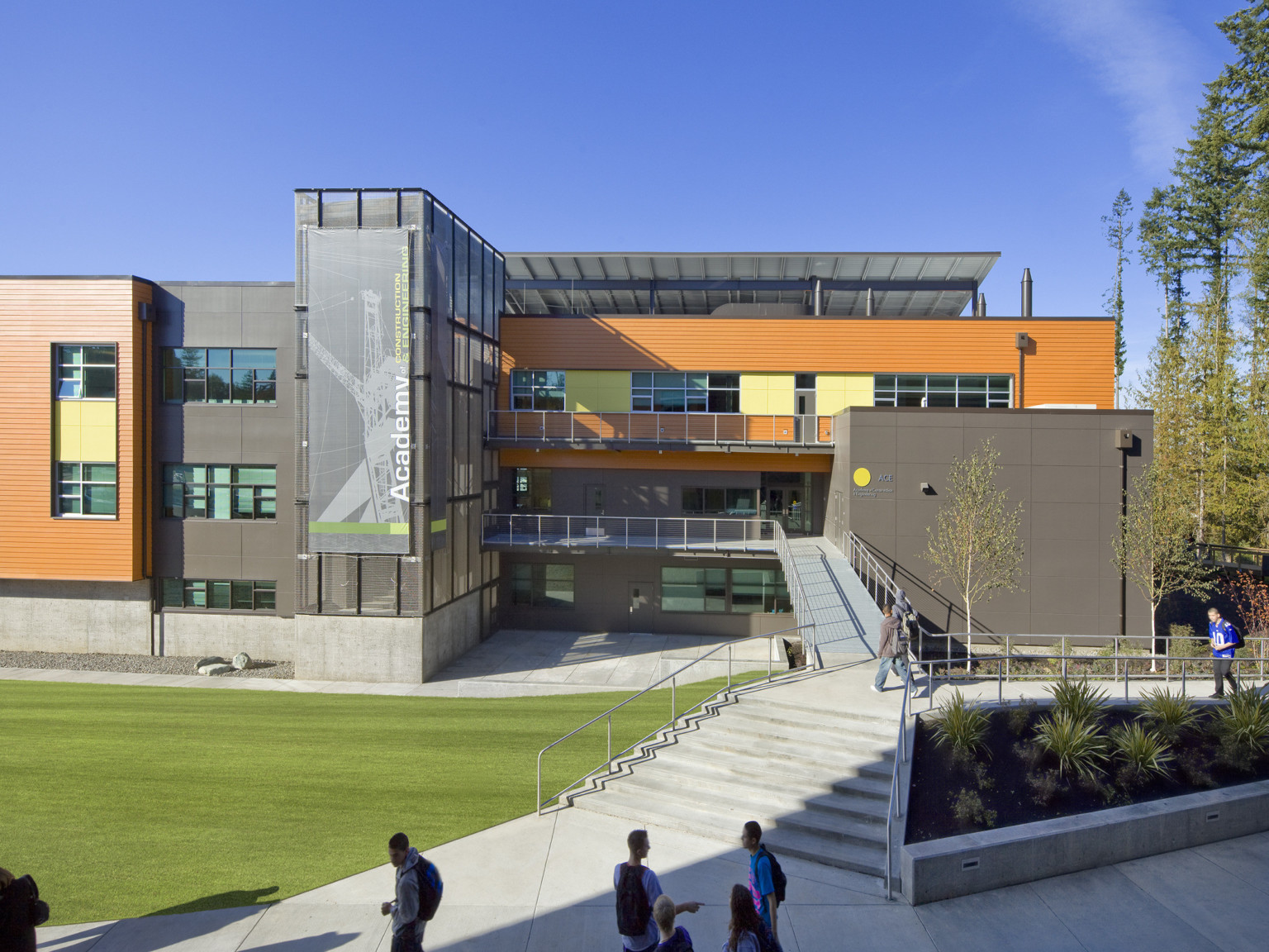 Concrete steps to L shaped ramp. Triple height glass tower extends from building, left between grey and orange facades