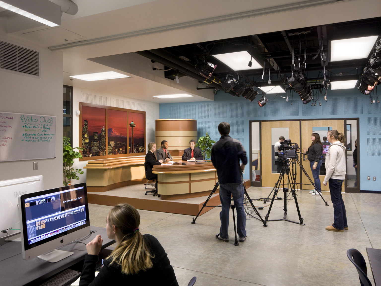 Broadcast classroom with cameras and lighting equipment pointed at wood news desk. Computer with editing equipment to left.