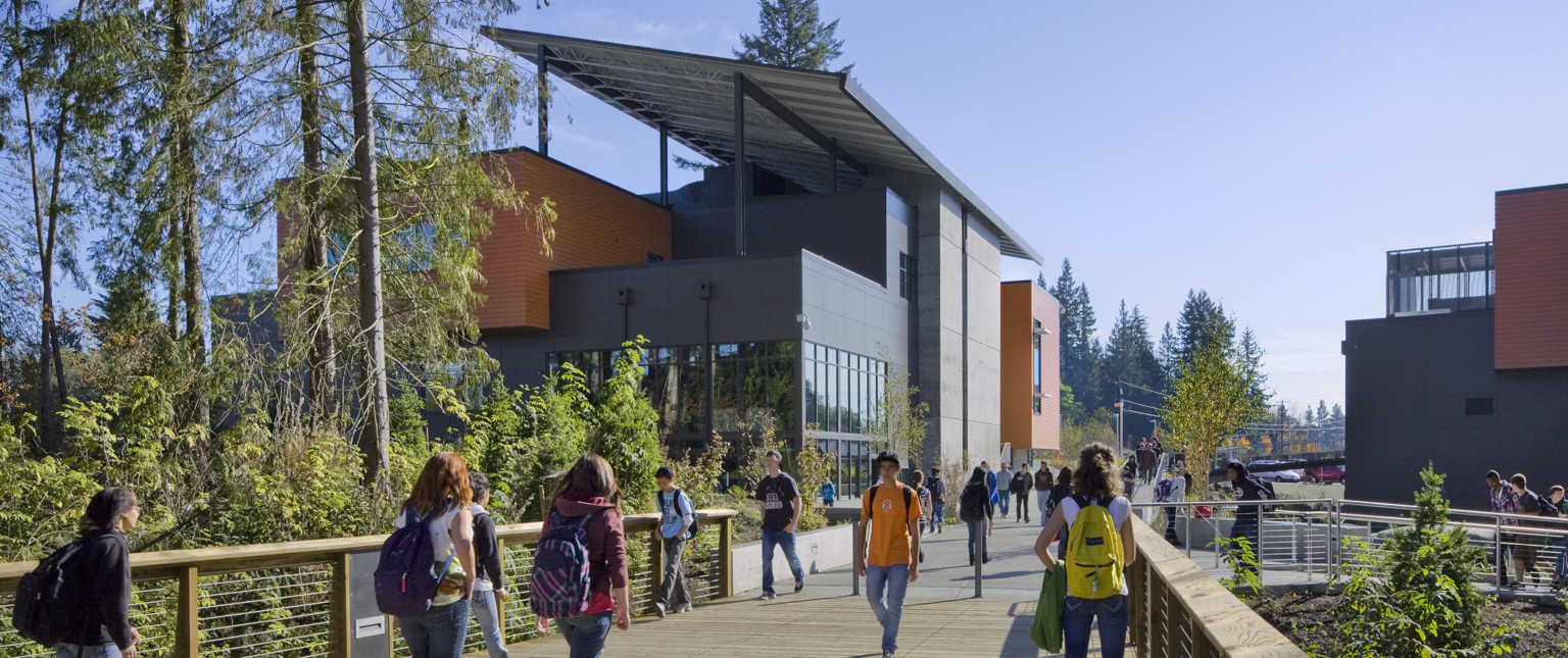 Wood bridge over trees looking to grey 3 story building with orange panel accents and large roof panel angled across top