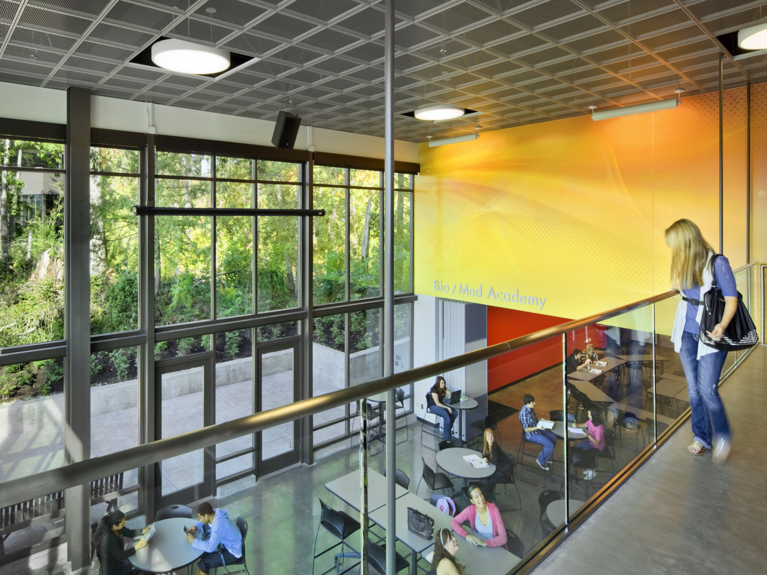 2nd floor walkway in double height atrium with windows looking out to trees. Yellow accent wall to right over seating area