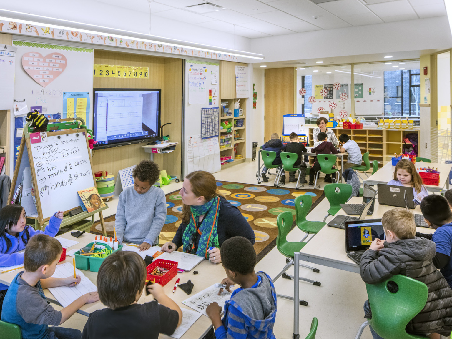 A classroom with white walls and light wood details. Alphabet mat at center surrounded by tables and green chairs