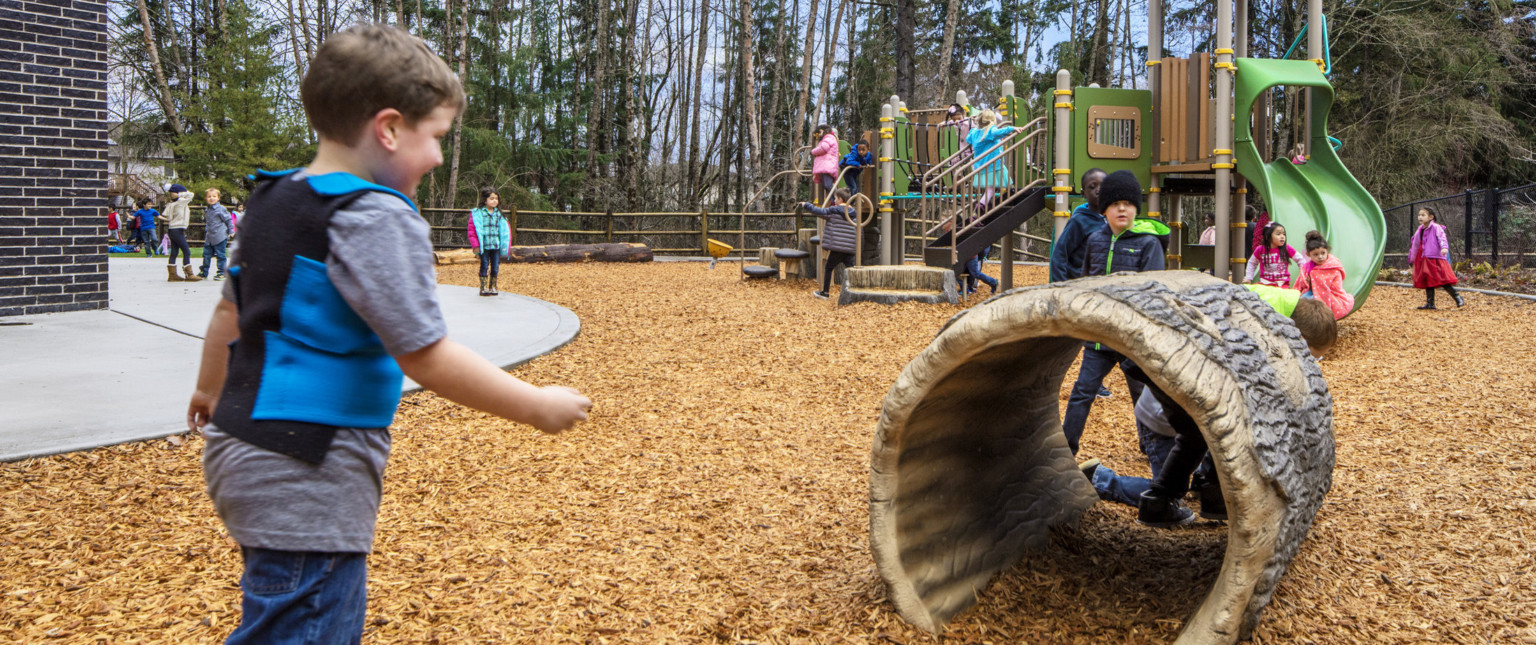 Playground equipment with slide and tree log tunnel on yellow rubber mulch. School building's dark brick is visible left