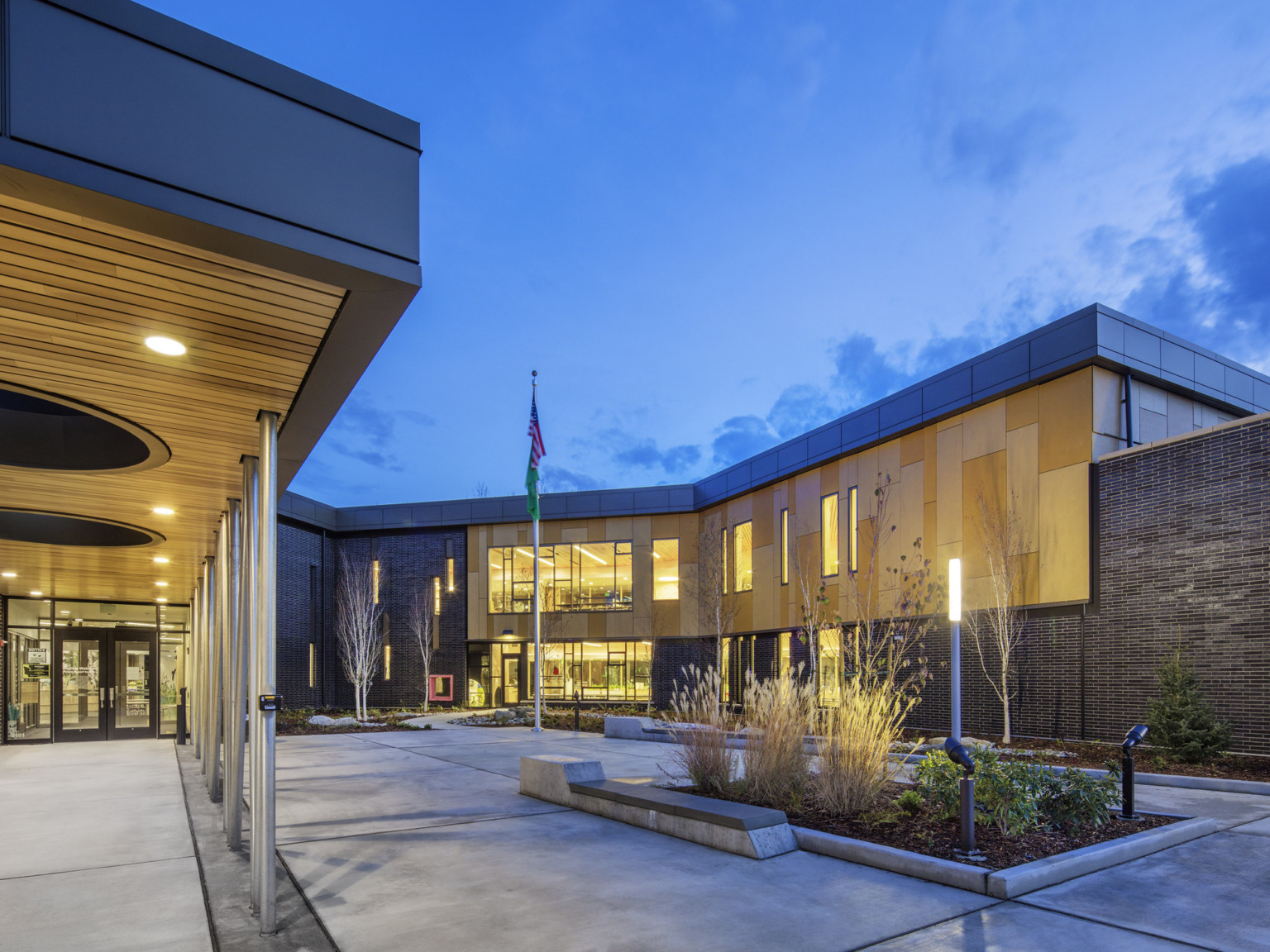 Front entry to building with patio shelter covering walkway. Right, a building wrapped courtyard with planters and flag