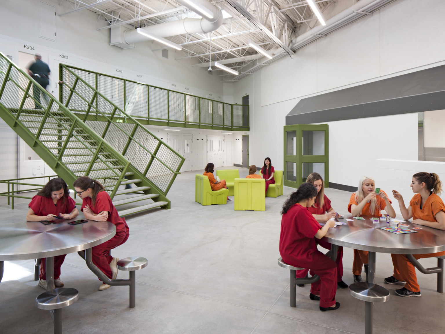 2 levels of cells connected by green stairs and railings leading to central area with green chairs, metal tables with chairs