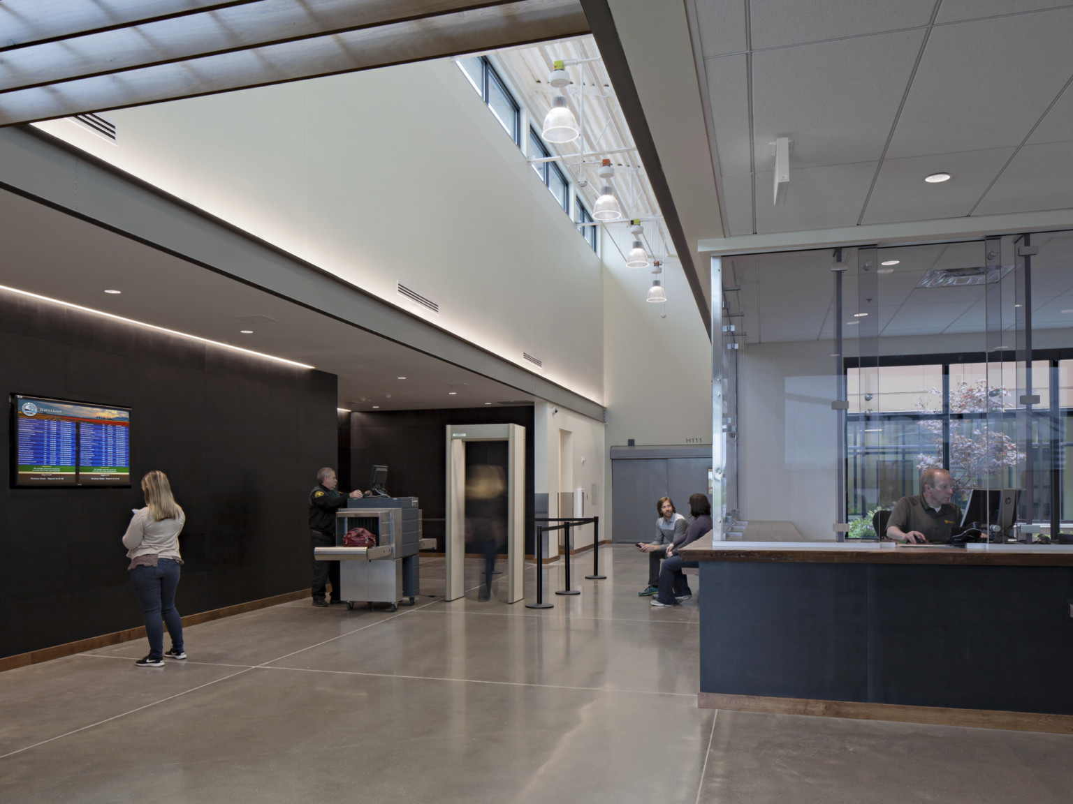 Double height hallway with recessed side walls. Glassed in reception desk, right, and metal detector, left. Lights hang above