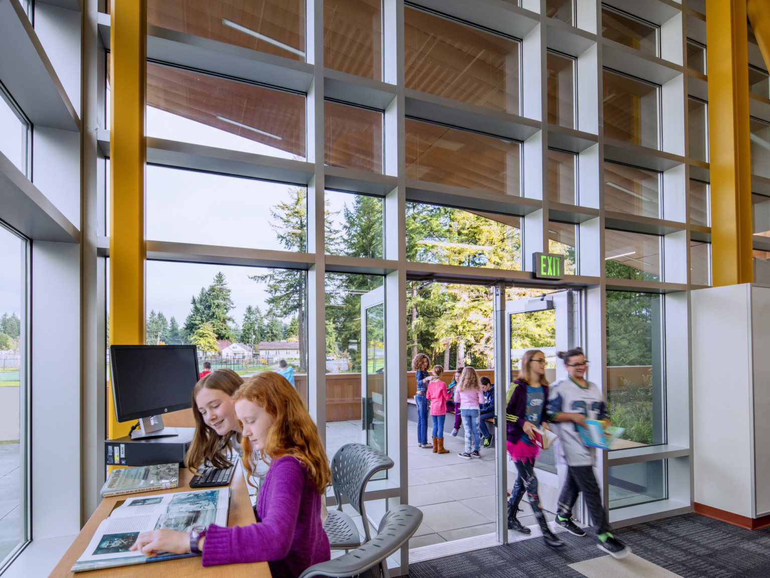 Corner of double height glass room with desk and computer. Windows have silver seams and double doors. Yellow column accents