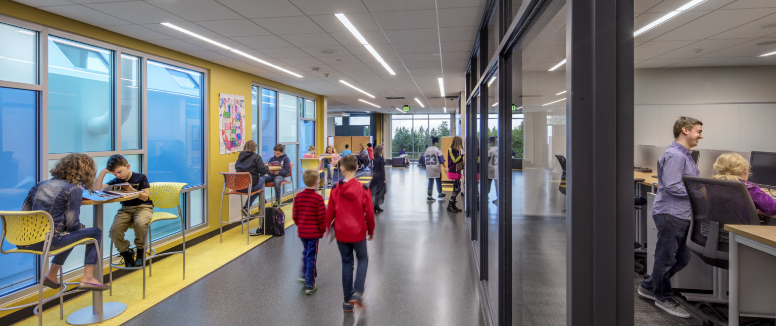 Interior hallway with yellow wall, left, with blue translucent windows. Glass walls into classrooms, right. High top tables