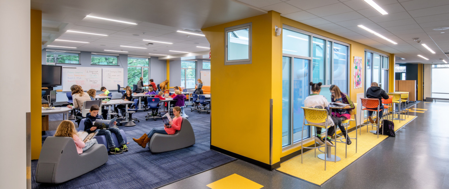 Hallway with high top tables and chairs separated from flexible and ergonomic seating by yellow wall with sheer blue windows