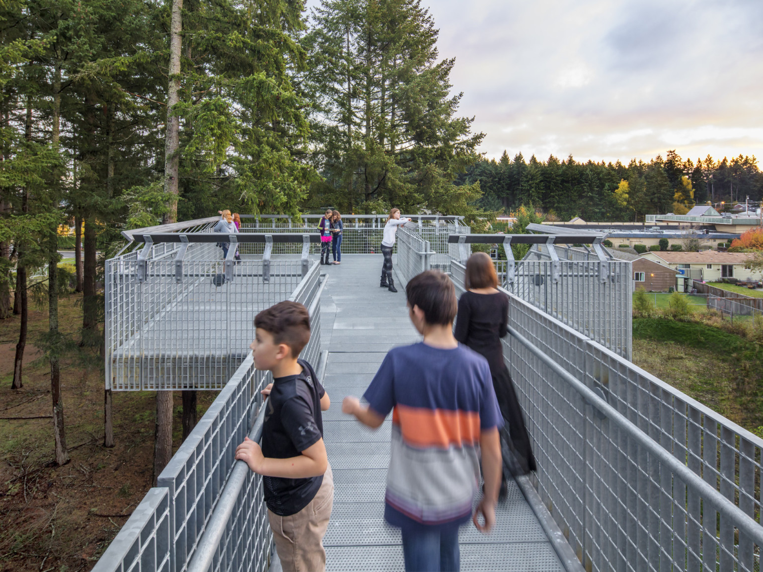 Silver metal exterior elevated walkway and balcony looks out to woods, left, and buildings, right