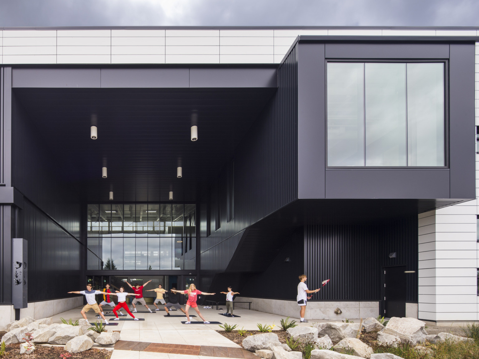 Exterior view, atrium is recess and patio is covered. Right, the building extends in a black tunnel with large window at end