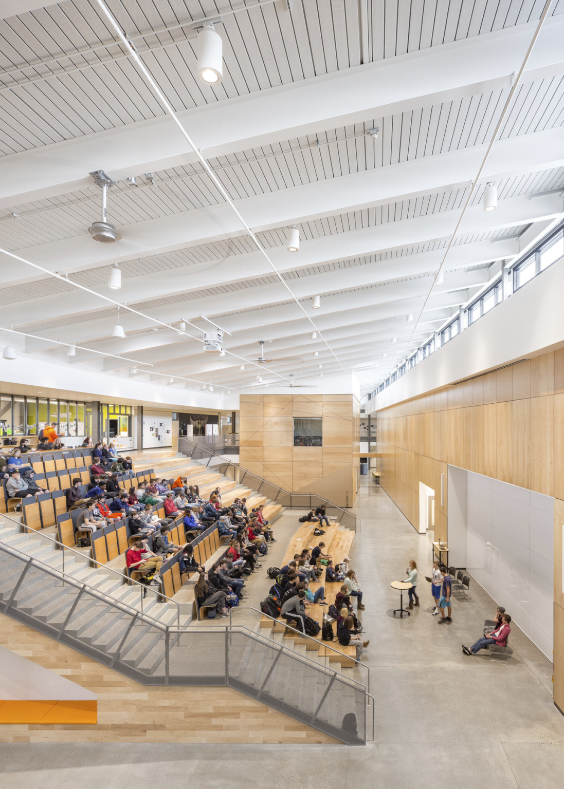 Wood paneled room with bleachers and folding theater seats under angled white roof. Wooden box structure beyond with window