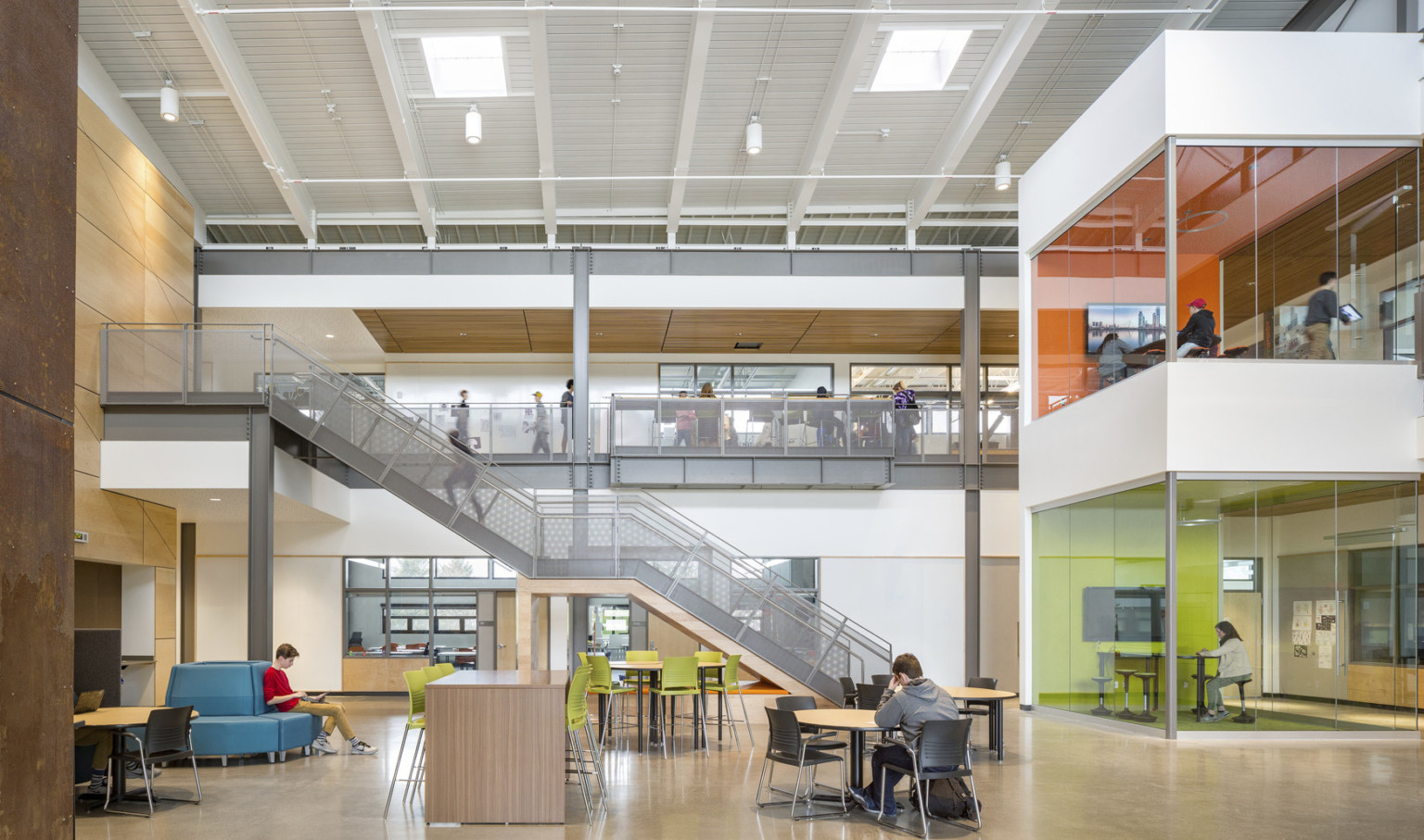 Atrium in Discovery High School with double height ceiling and 2 levels of colorful glass learning pods. Grey stairs, left