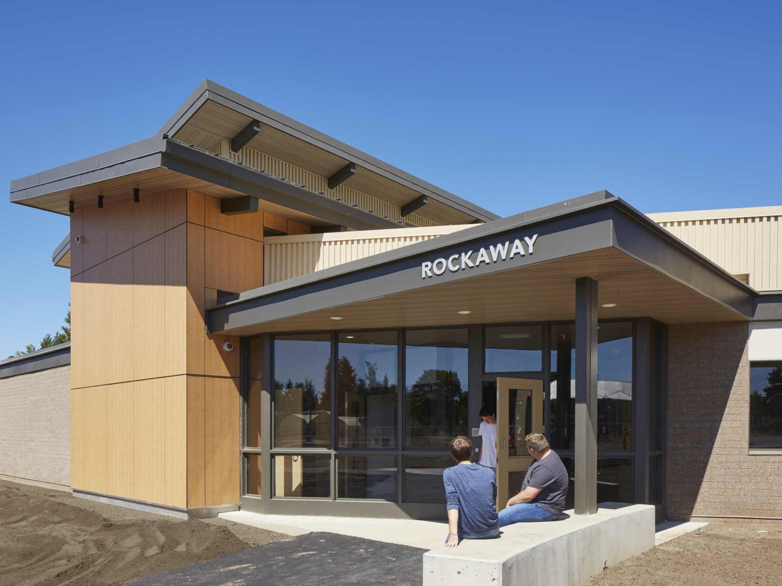 Exterior view of building entry with pointed overhang with wood panel base supported by black column anchored in cement bench